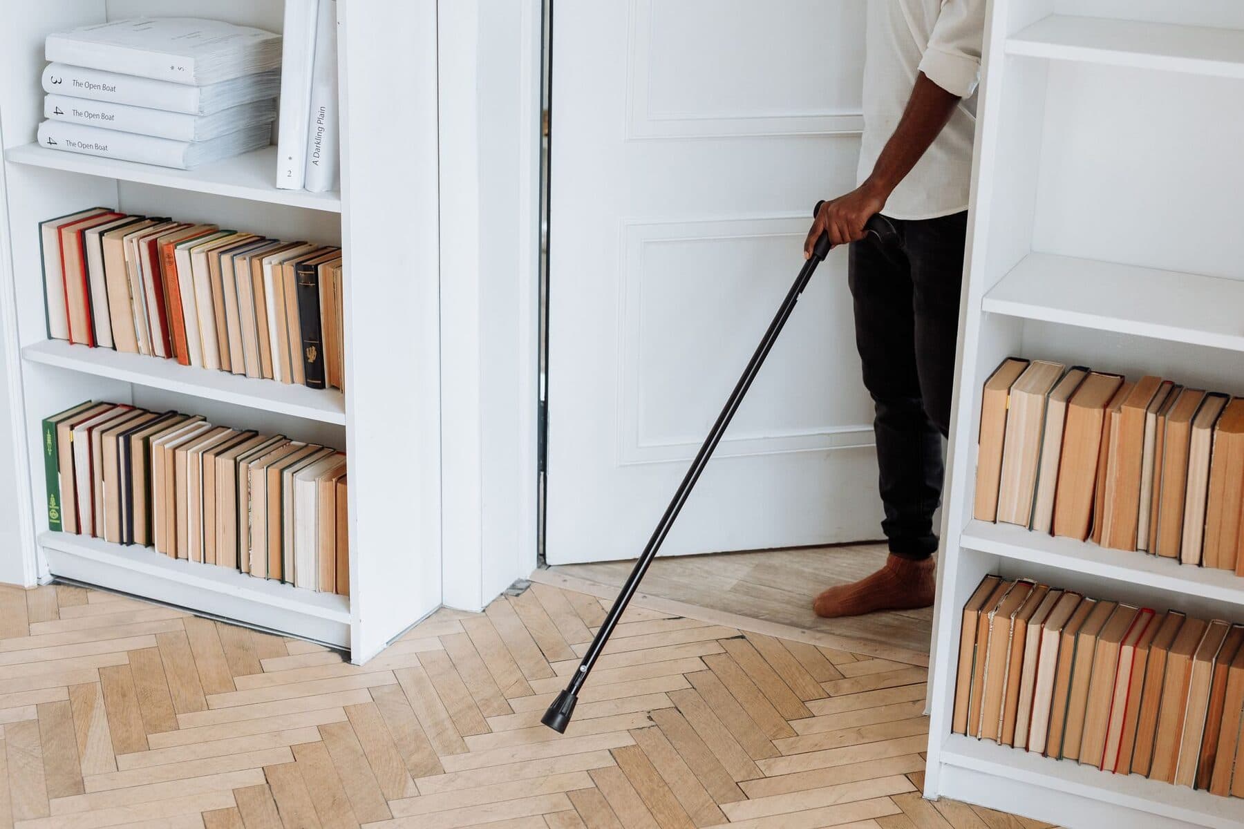 Un bastone da passeggio nero tenuto da una persona di spalle, accanto a una libreria bianca in una stanza luminosa