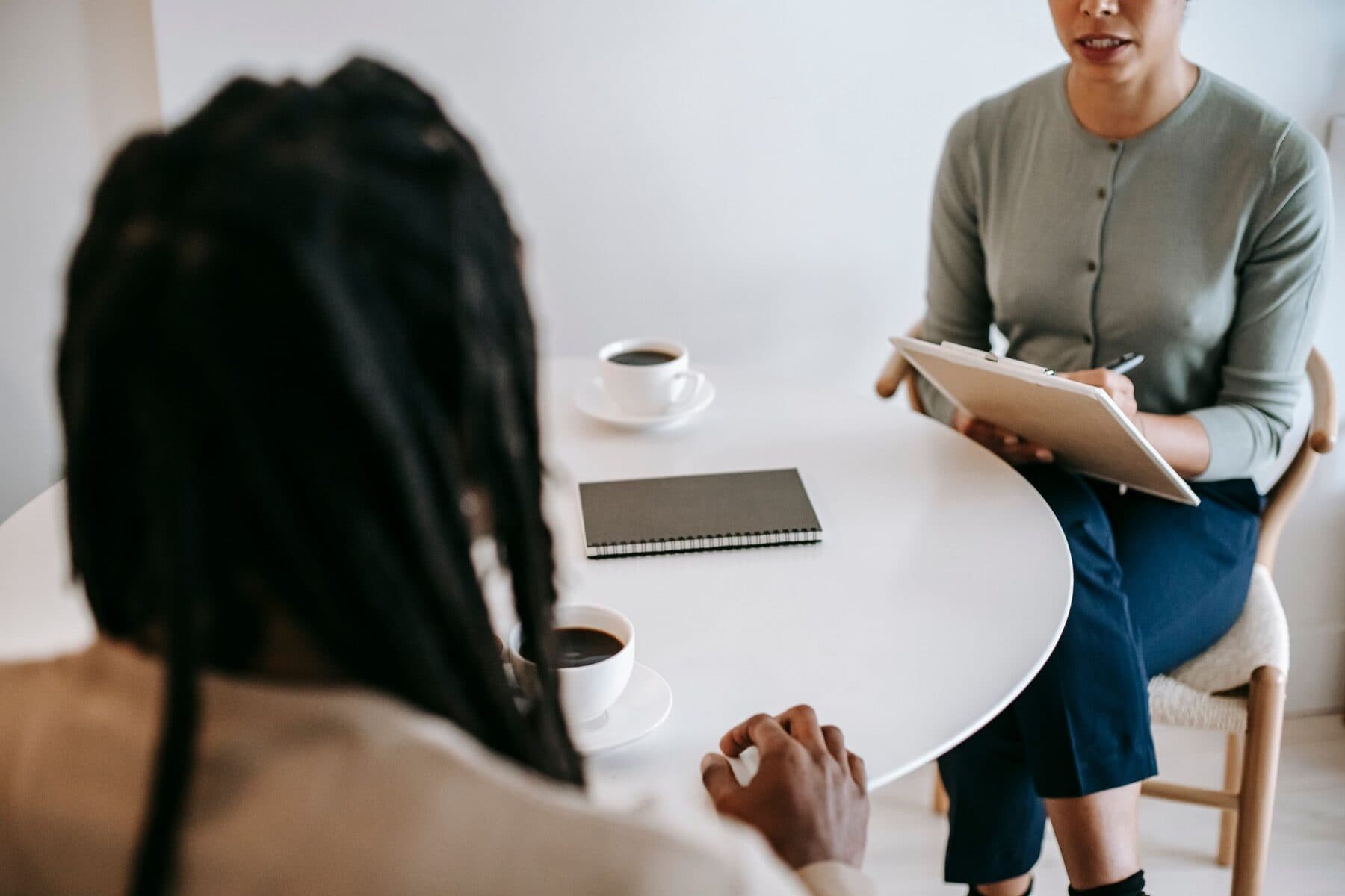 Una consulente con la tavoletta appunti parla seduta a un tavolo bianco con una cliente di spalle, accanto a un quaderno e a una tazza di caffè