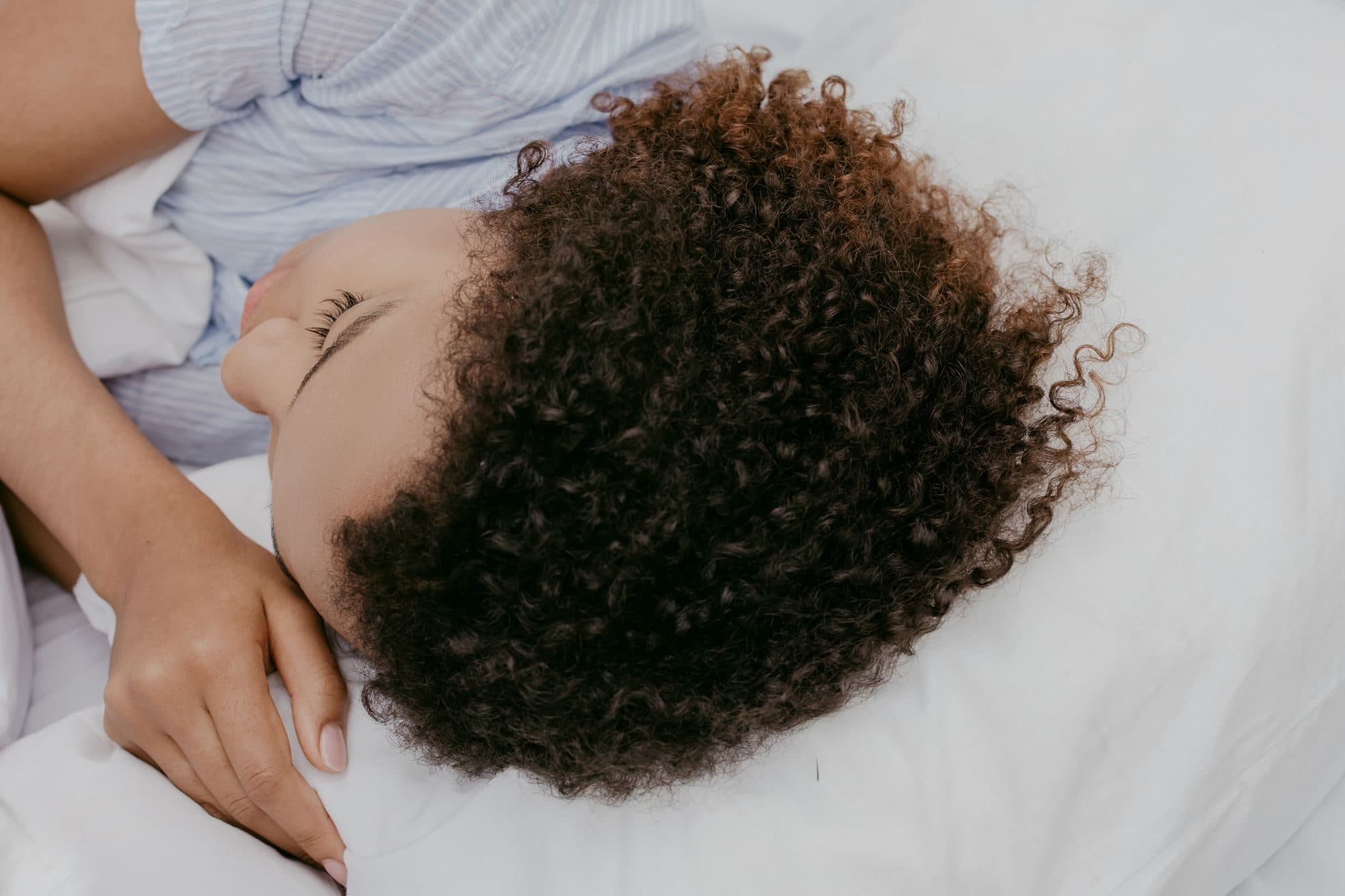 Una persona si riposa di fianco con i capelli ricci sul cuscino bianco, in una luce morbida e chiara